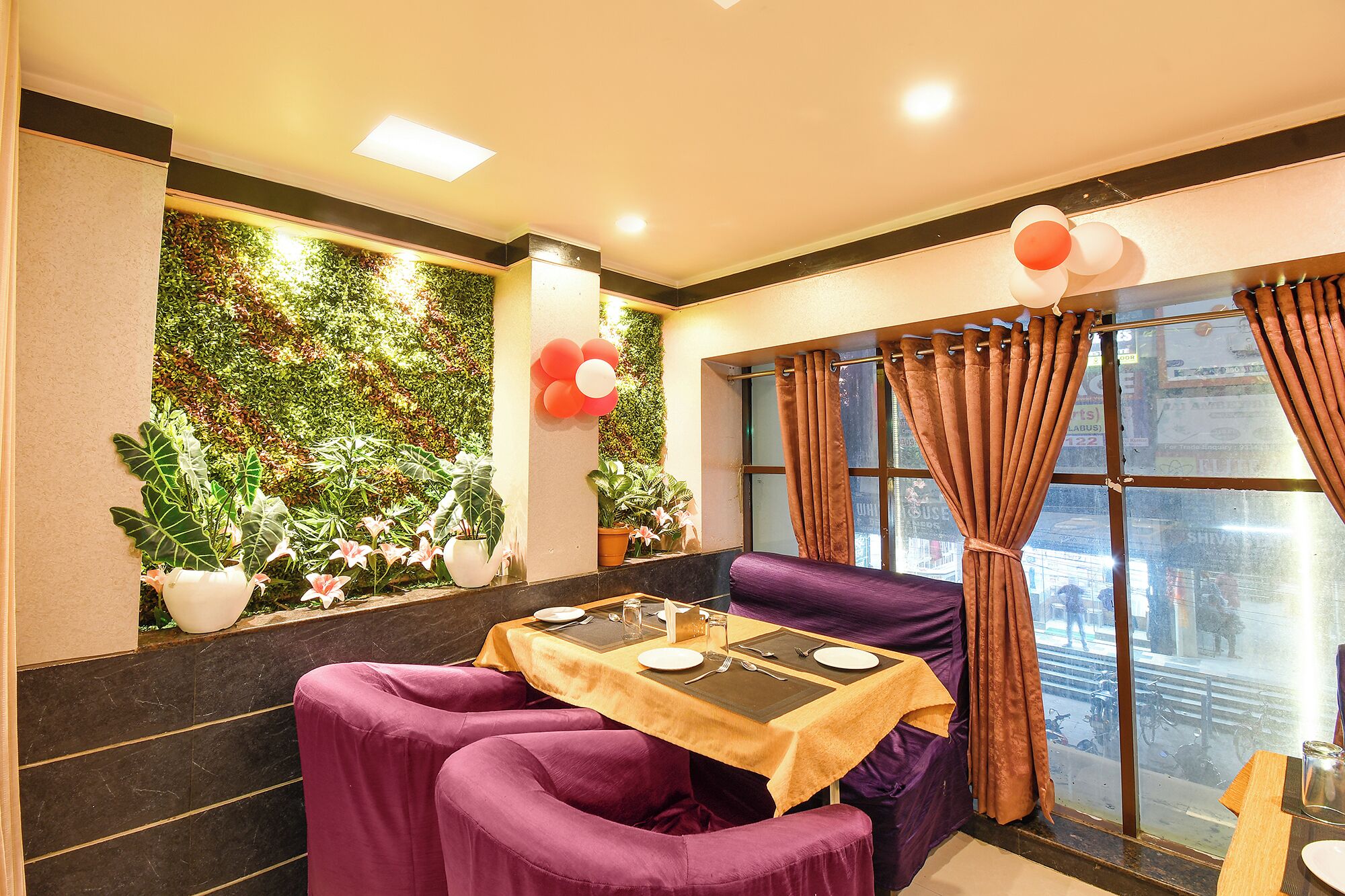 A dining area with a table set for two, purple upholstered chairs, a green wall with plants, and a window with curtains.