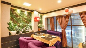 A dining area with a table set for two, purple upholstered chairs, a green wall with plants, and a window with curtains. - Fabhotel Raj Matrya Inn (Patna)