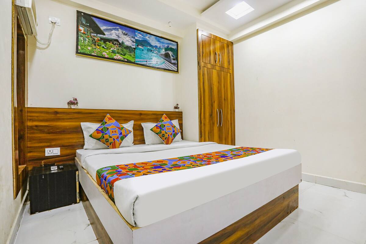 A bedroom with a wooden headboard, a bed with a colorful geometric-patterned bedspread, a nightstand with a black suitcase, and a large wardrobe.
