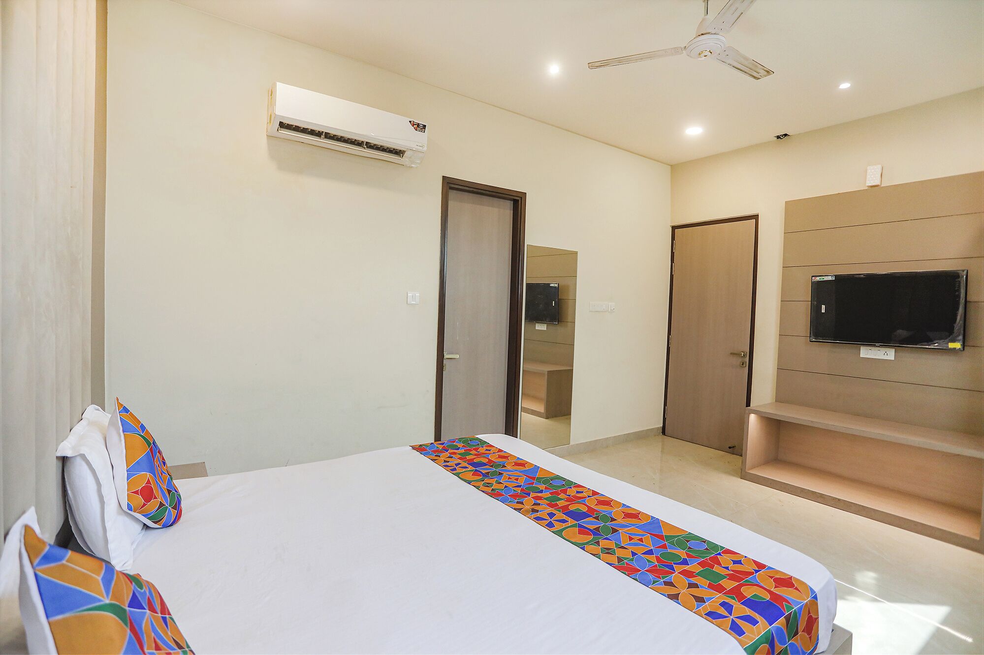 A hotel room with a bed, a TV mounted on the wall, an air conditioning unit, and a ceiling fan.