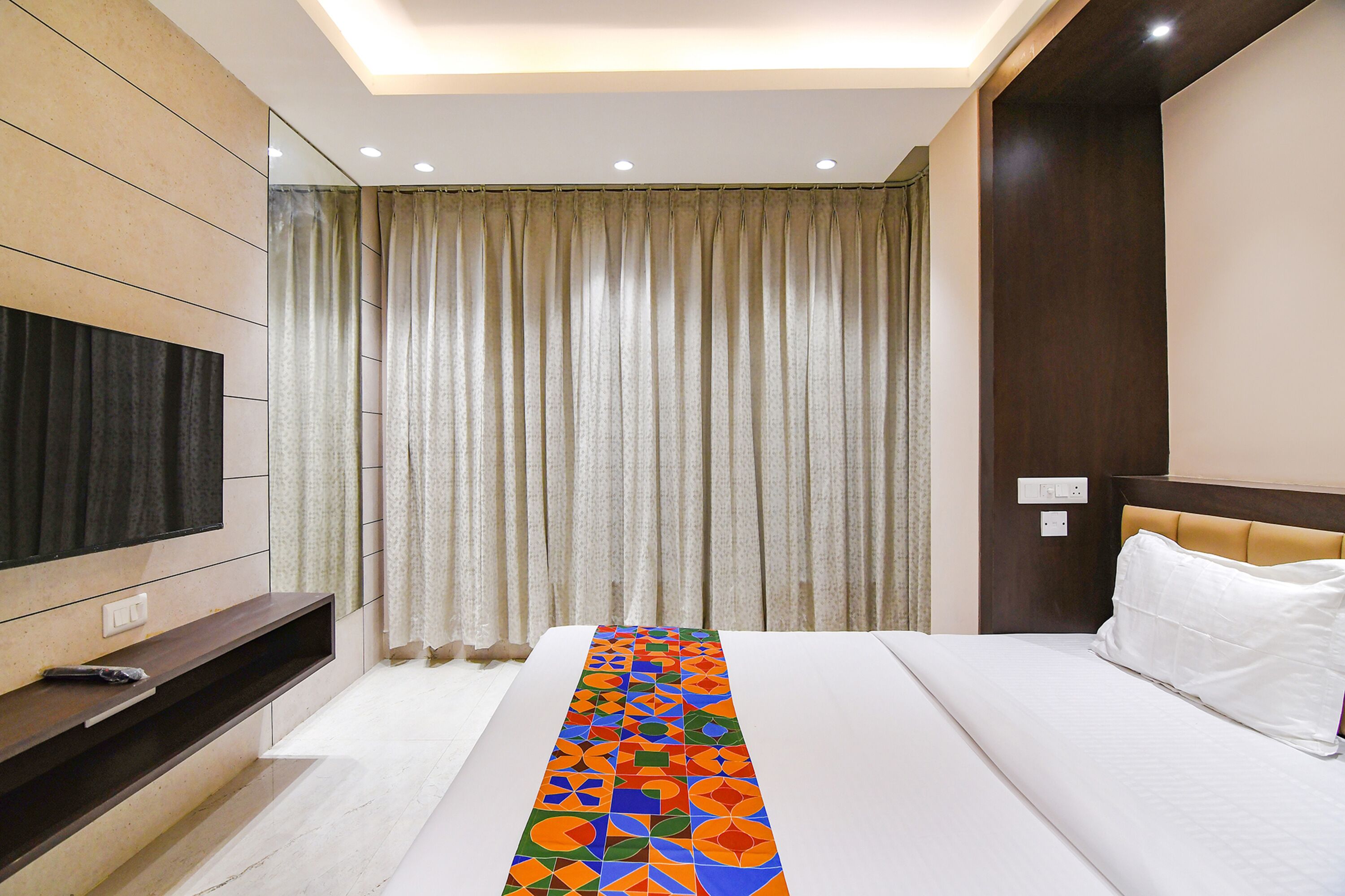 A hotel room with a bed, a TV mounted on the wall, a long decorative pillow, and a window with curtains.
