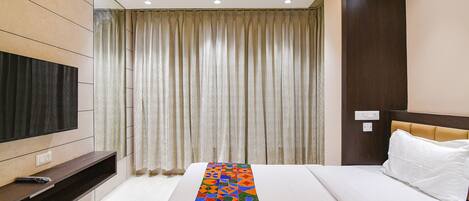 A hotel room with a bed, a TV mounted on the wall, a long decorative pillow, and a window with curtains.