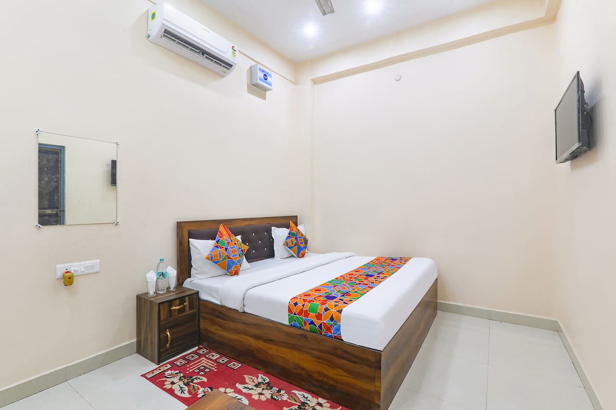 A bedroom with a wooden bed, white bedding, a colorful geometric throw, a small bedside table with a bottle, a red floral rug, an air conditioning unit, a wall-mounted TV, and a mirror.