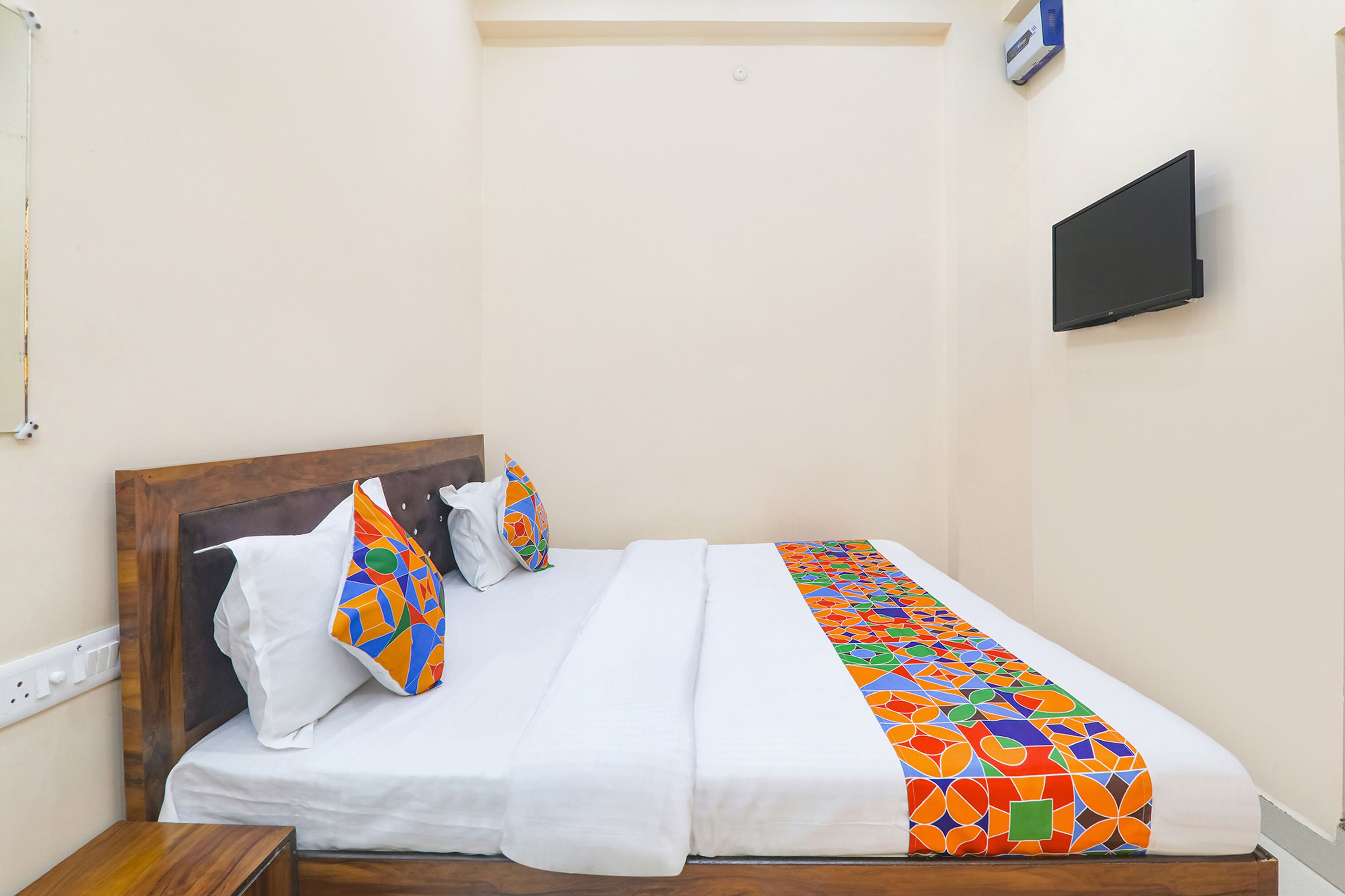 A hotel room with a wooden headboard, a white bedspread, colorful pillows, and a TV mounted on the wall.