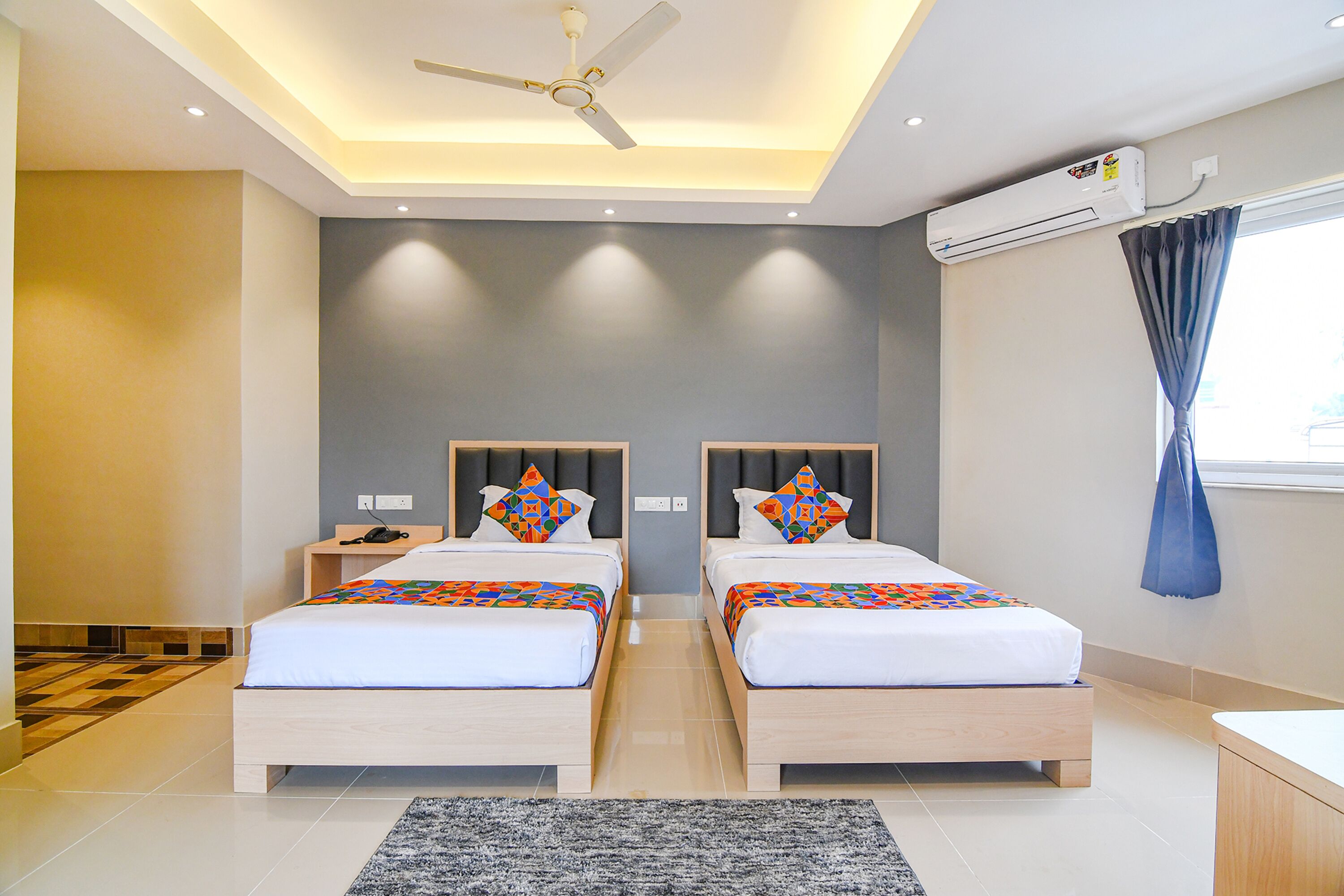 A modern hotel room with two beds, a ceiling fan, recessed lighting, an air conditioning unit, and a large window with curtains.