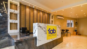 Uma área de recepção de hotel com balcão de mármore, uma placa 'Tab Hotels', uma câmera de segurança e uma janela com persianas.