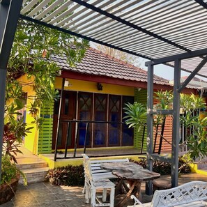 A covered patio area with a wooden table and benches, surrounded by potted plants and a colorful building with a tiled roof. - Baan Siri Mukdahan (Mukdahan)