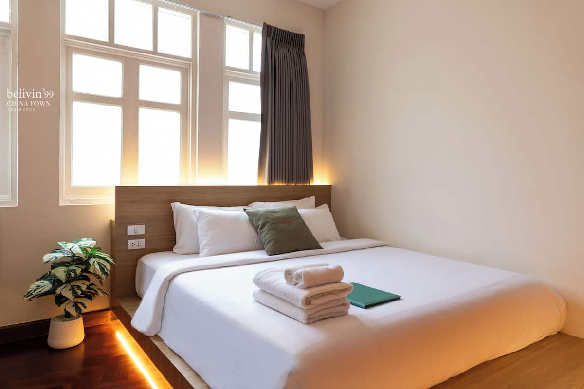 Een net opgemaakt bed met witte lakens, een groen kussen en een opgevouwen handdoek. Op de vloer naast het bed staat een potplant.
