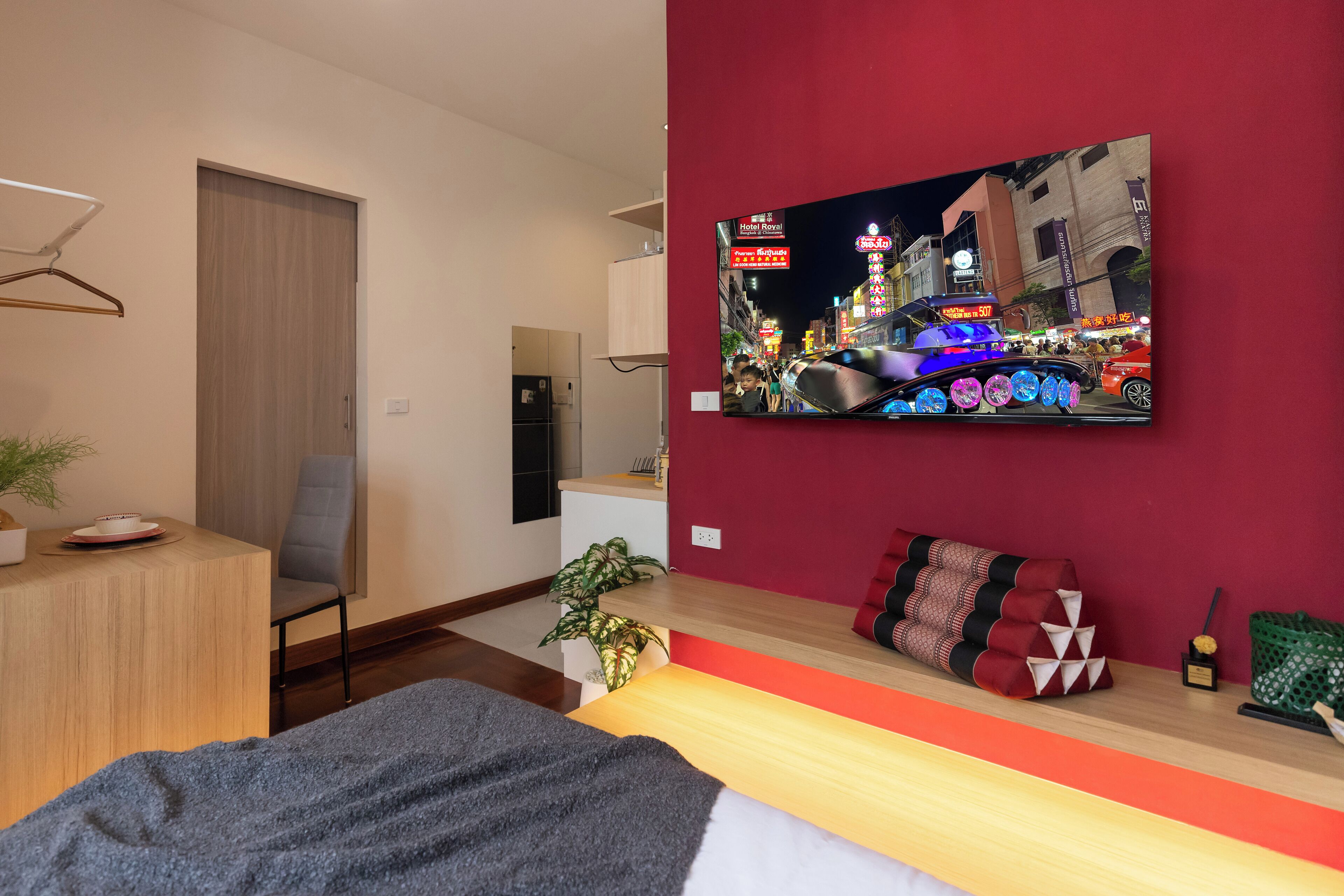 A modern hotel room with a bed, a desk, a chair, a TV on the wall, and a red accent wall.