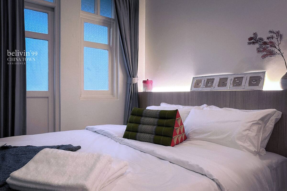 Een net opgemaakt bed met witte lakens, een grijze deken en decoratieve kussens. Er is een raam met gordijnen, een wandlamp en een kleine plant op het nachtkastje.