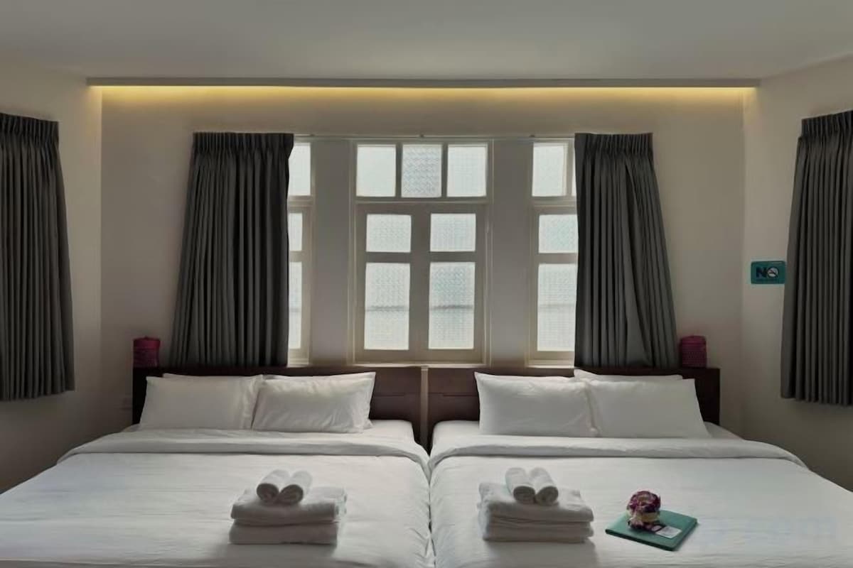 A hotel room with two beds, a large window with curtains, and a bedside table with a lamp.