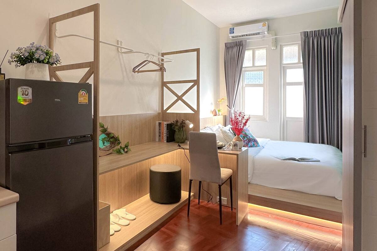 Een kleine hotelkamer met een bed, een bureau, een stoel, een kitchenette en een raam met gordijnen.