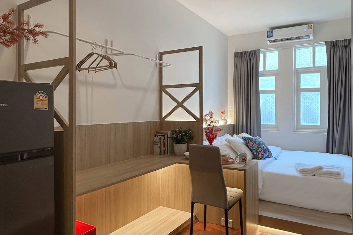 Een knus hotelkamer met een bed, een bureau met stoel, een klein tafeltje, een koelkast en een raam met gordijnen.