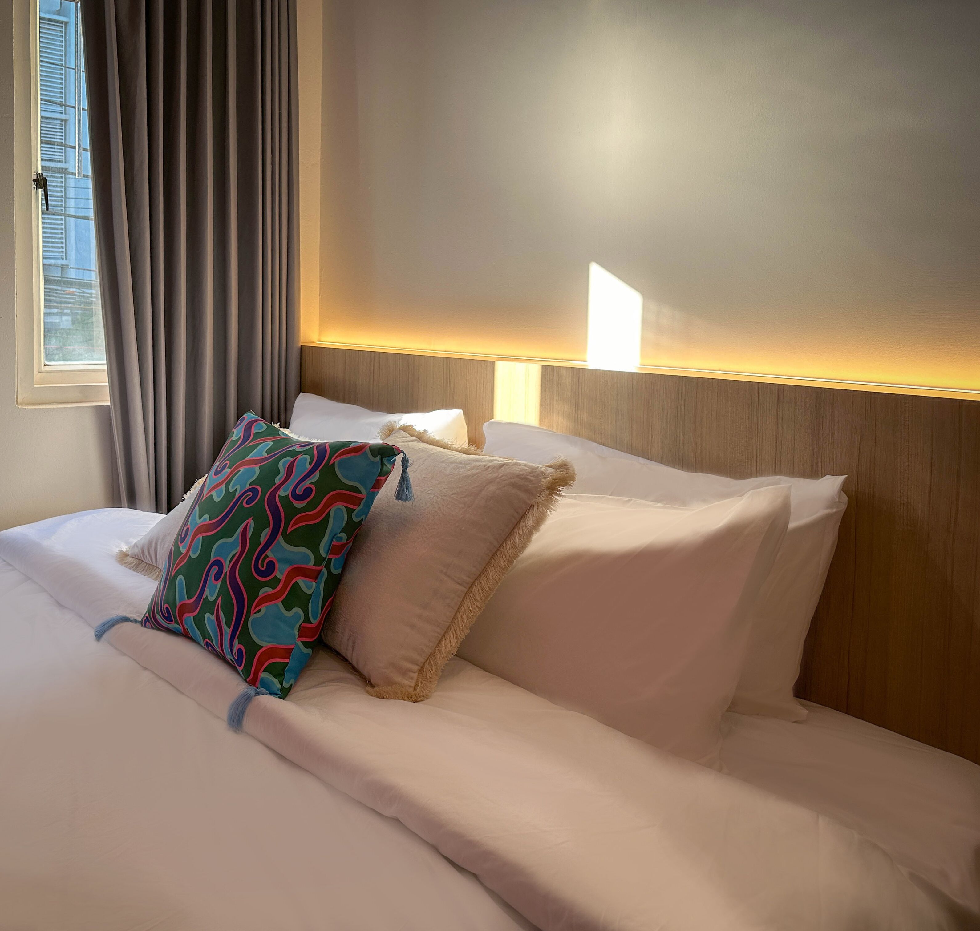 A hotel room with a neatly made bed, a window with curtains, and a wall-mounted light fixture.