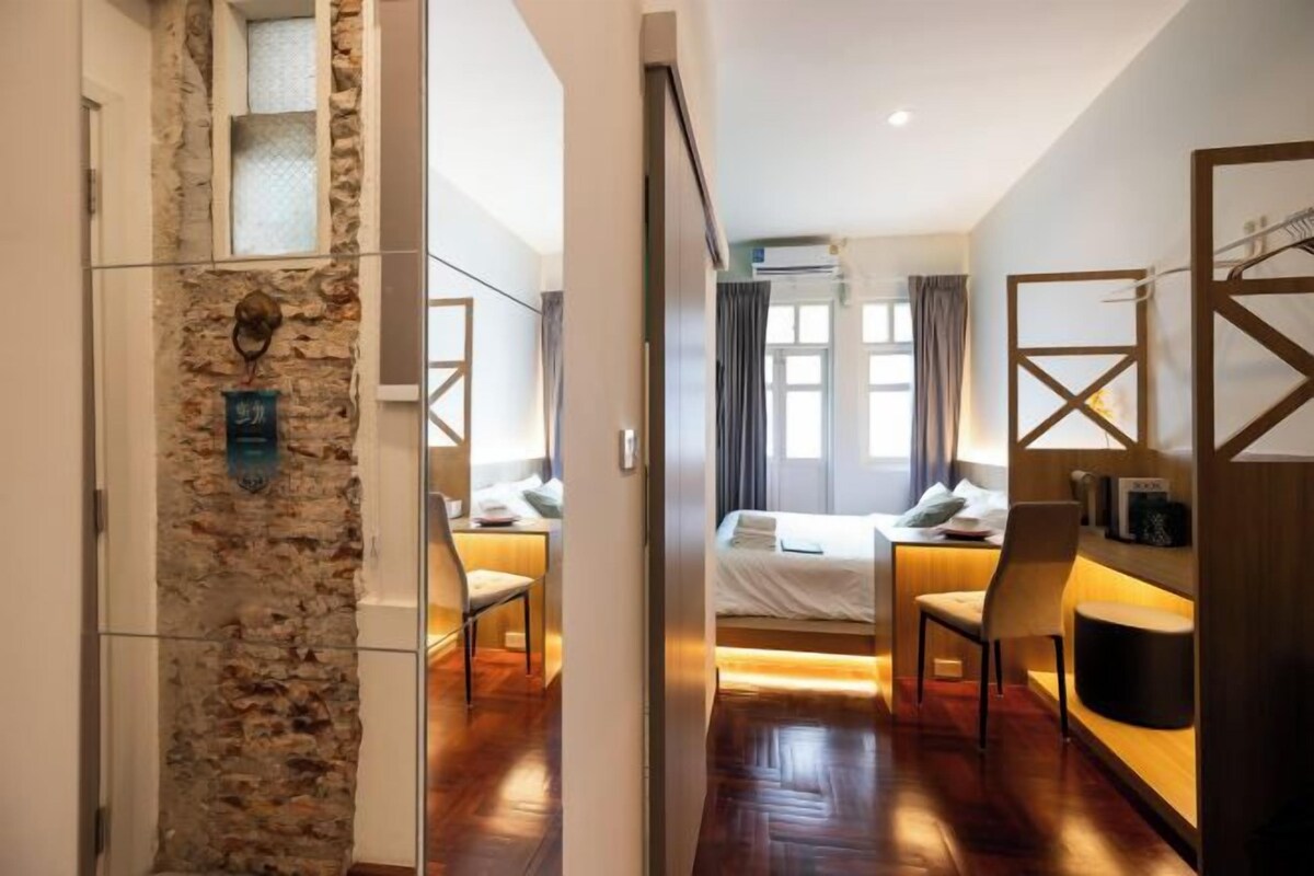 Quarto de hotel com parede de pedra, cama com lençóis brancos, escrivaninha com cadeira e armário de madeira.