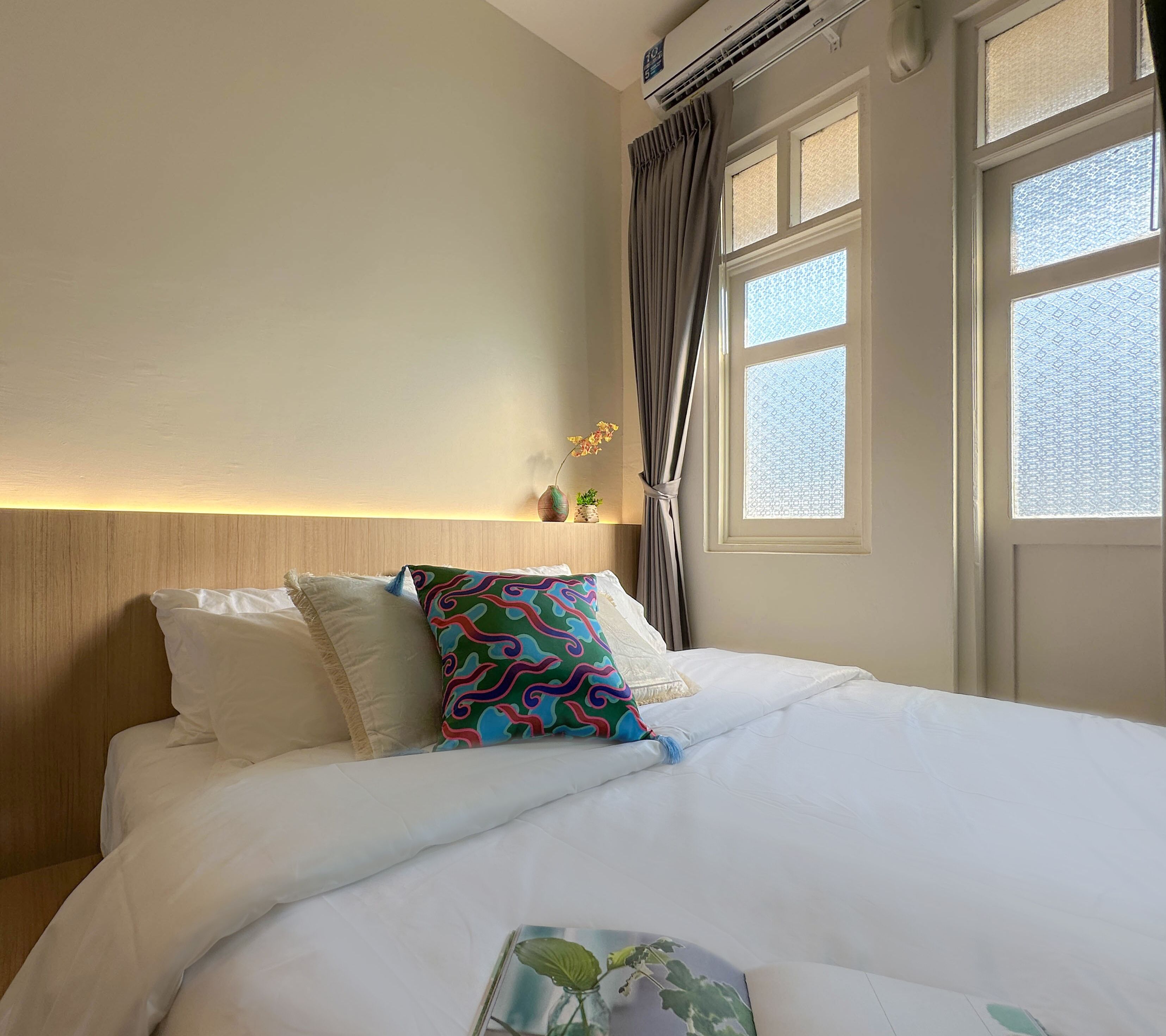 A neatly made bed with white bedding and colorful pillows, a bedside table with a vase and a small plant, and a window with patterned curtains.