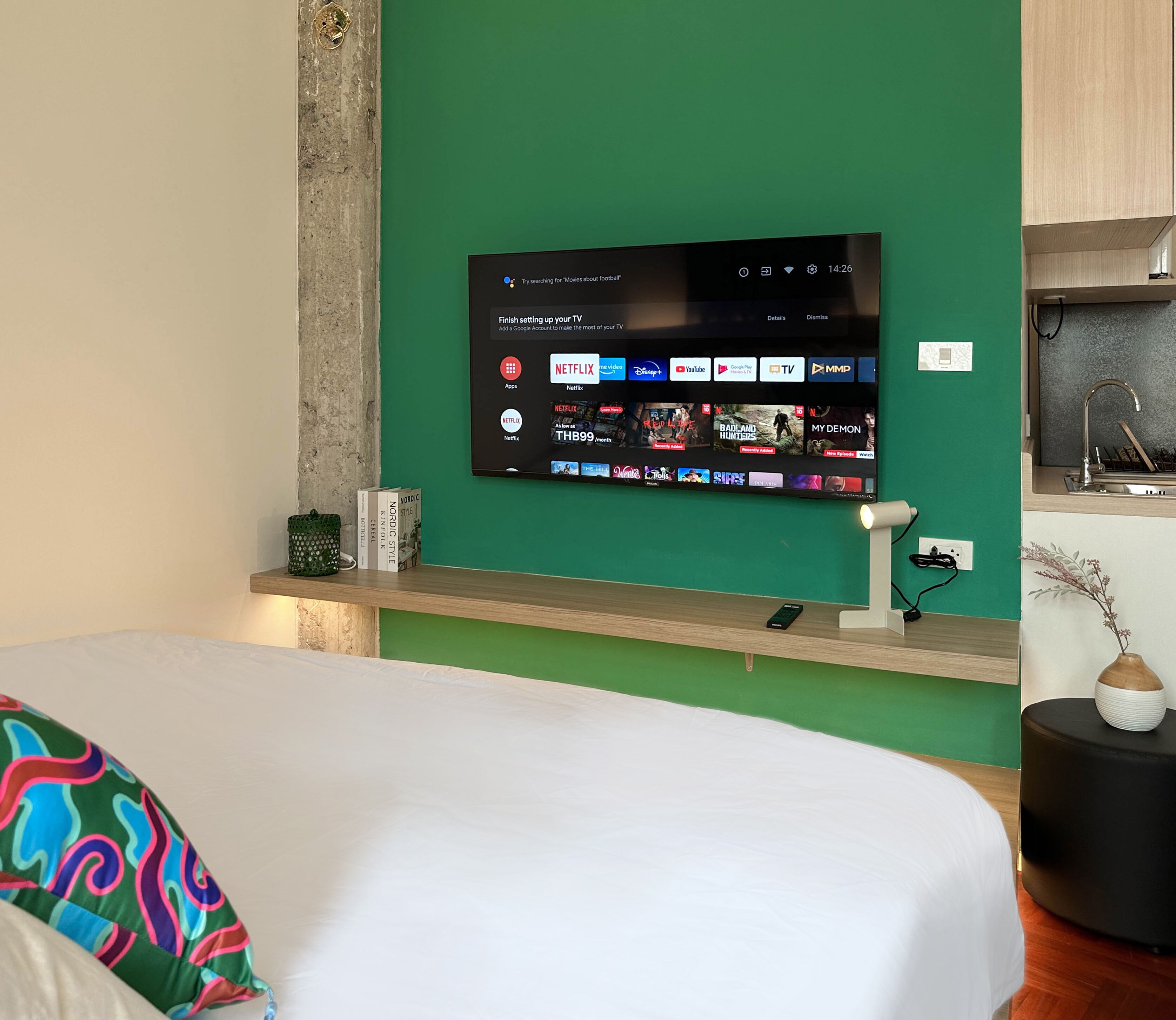A modern hotel room with a large flat-screen TV mounted on a green wall, a wooden bedside table with a remote control, and a bed with a colorful pillow.