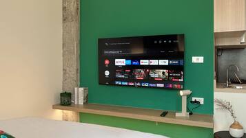 A modern hotel room with a large flat-screen TV mounted on a green wall, a wooden bedside table with a remote control, and a bed with a colorful pillow.