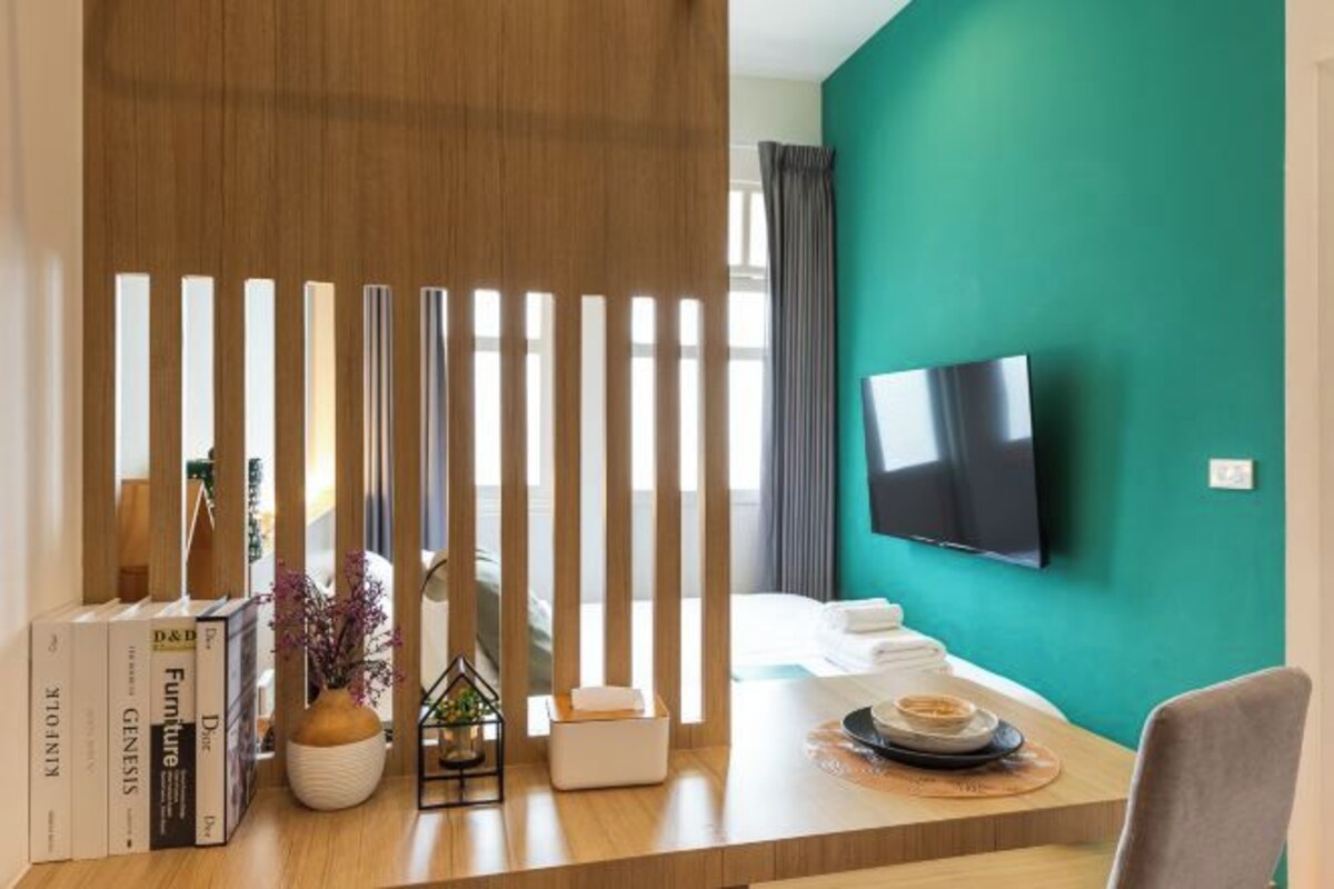 A hotel room with a teal accent wall, a flat-screen TV mounted on the wall, a wooden desk with books, a vase with flowers, and a bed with white linens.