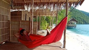 Iemand ontspant in een rode hangmat op een houten terras, met uitzicht op een strand en een huis op palen in de verte.