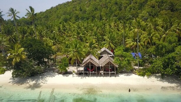 Een resort aan het strand met bungalows met rieten daken, palmbomen en helder turquoise water.