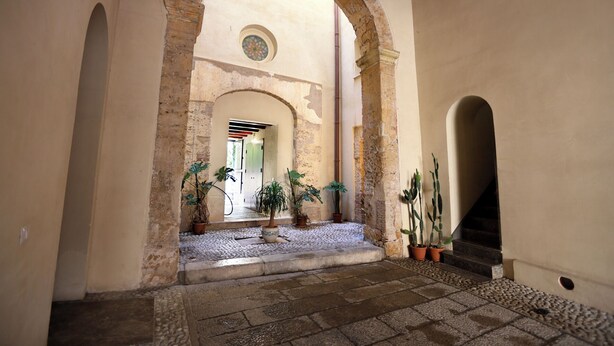 An arched corridor with stone flooring, potted plants, and a view into a room with a door. - Elegant apartment in historic building in the heart of the Kalsa Palermo neighborhood. (Palermo)
