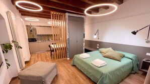 A compact bedroom with a bed, bedside tables, a desk, and a kitchenette in the background. - Elegant apartment in historic building in the heart of the Kalsa Palermo neighborhood. (Palermo)
