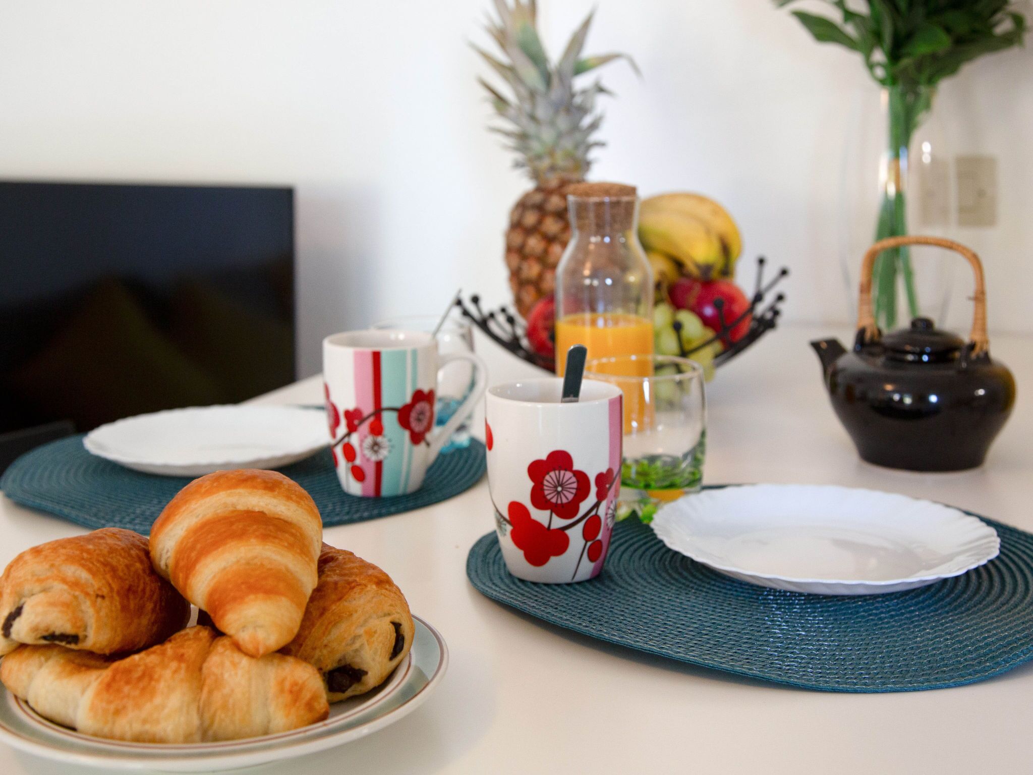Une table dressée pour le petit-déjeuner avec des croissants, un ananas, une bouteille de jus d’orange et une théière.