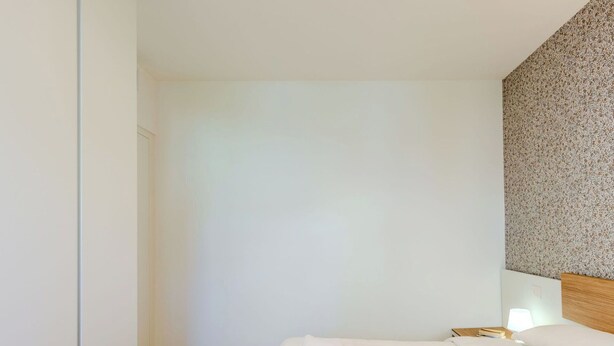 A hotel room with a wooden floor, a bed with white linens, a nightstand with a lamp, and a textured wall.