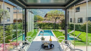 A glass-enclosed dining area with a pool view, outdoor seating, and a table set with a bottle and glasses.