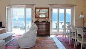 A room with a large window, a wooden dresser, a white bean bag, and a dining area with chairs. - Casa di CLO sea front stone property,private pool, Fiscardo 500 m (Borgo Fiskardo)