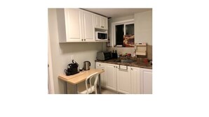A compact kitchen with white cabinets, a microwave, a coffee maker, and a small window with a view of a building. - Cozy One Bed Suite Near Beach (White Rock)
