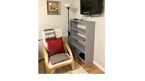 A small room with a wooden chair, a grey entertainment unit, a television, a floor lamp, and a framed picture on the wall. - Cozy One Bed Suite Near Beach (White Rock)
