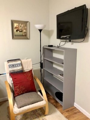Une petite pièce avec une chaise en bois, un meuble de rangement gris, un téléviseur, une lampe sur pied et un tableau encadré accroché au mur.