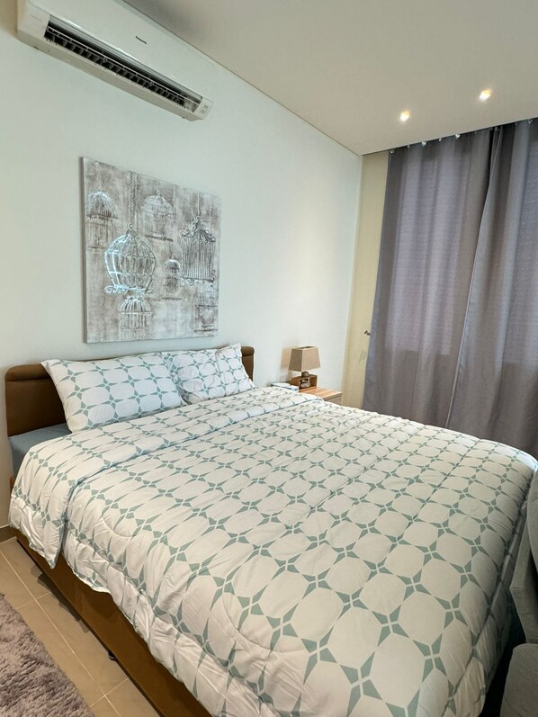 A bedroom with a large bed, a wall-mounted air conditioner, a nightstand with a lamp, and a window with curtains. - Azaiba Villas (Muscat)