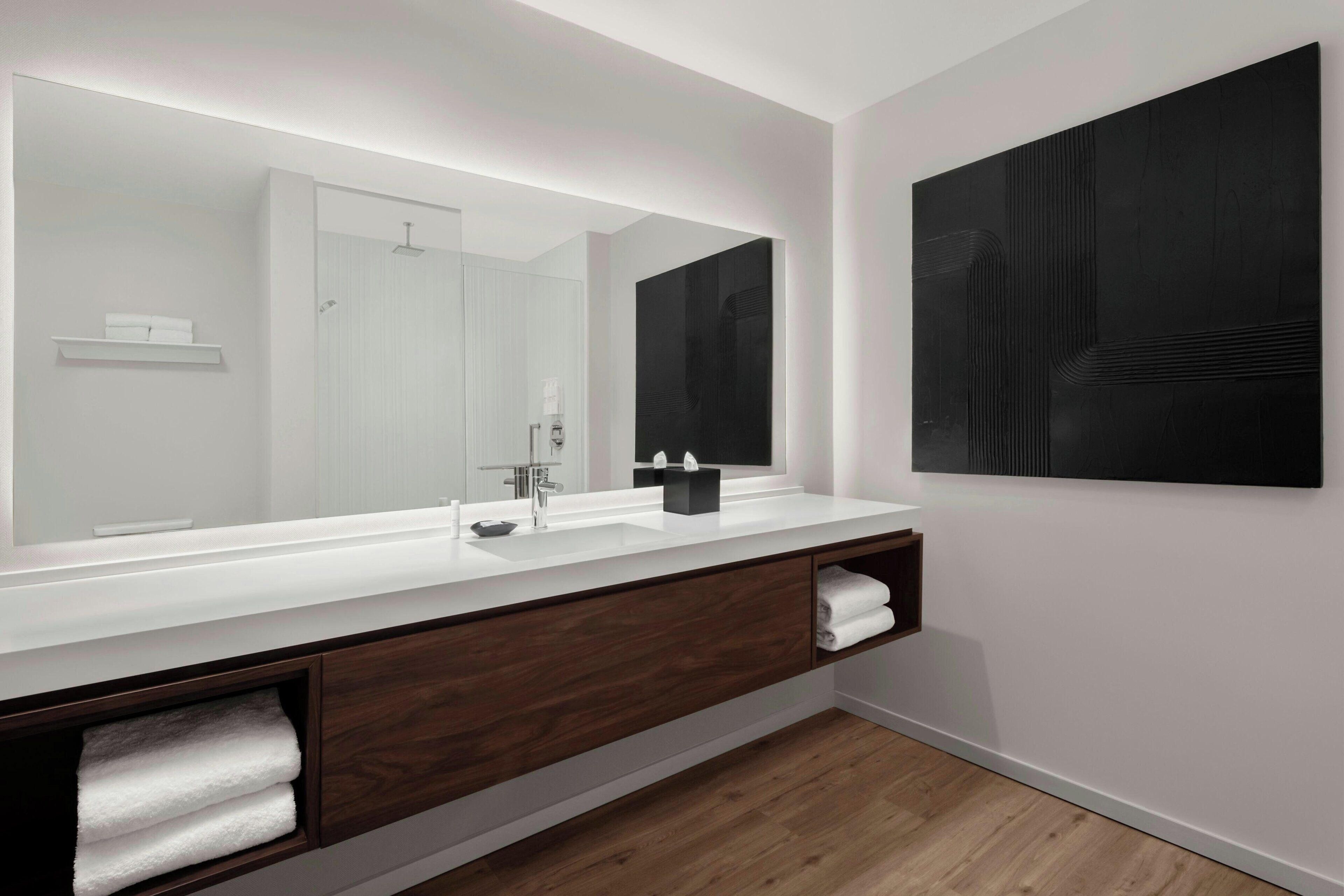A modern bathroom with a large mirror, white countertop, dark wood vanity, and a black abstract artwork on the wall.