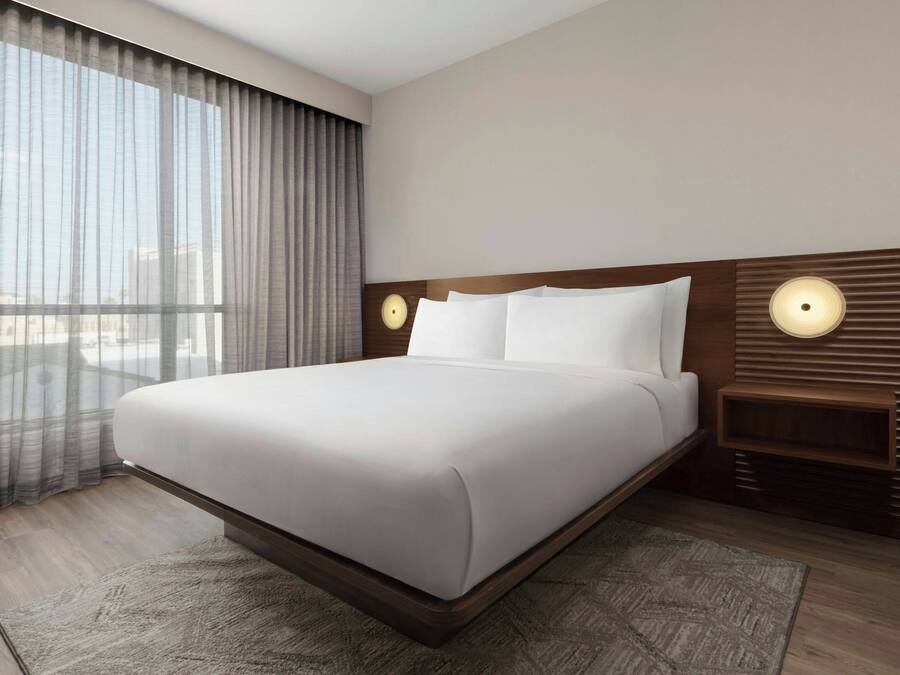 A modern hotel room with a large bed, two bedside lamps, a wooden headboard, and a view through sheer curtains.