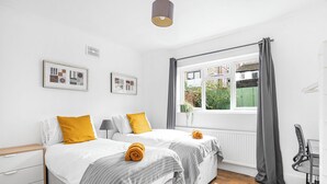 A modern bedroom with two beds, a desk, and a window with curtains. - Urban Haven (London)