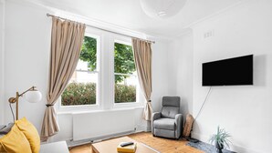 A living room with a sofa, a coffee table, a grey armchair, a flat-screen TV, and a window with curtains. - Urban Haven (London)