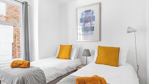 A hotel room with two beds, a window with curtains, a lamp, and a framed abstract artwork on the wall. - Urban Haven (London)