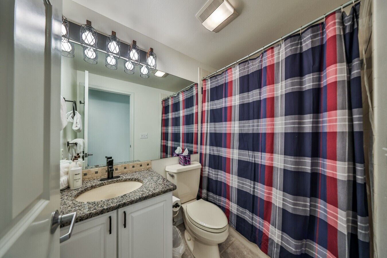Une salle de bain avec un lavabo sur un plan de travail en granit, des toilettes, un miroir avec éclairage et un rideau de douche à carreaux.