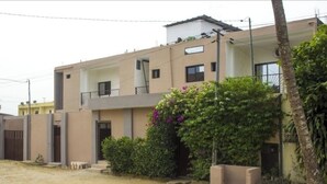 A two-story residential building with a balcony on the upper floor, a gated entrance, and a small garden area with flowering plants. - HIGH STANDING APARTMENT CI (Abidjan)