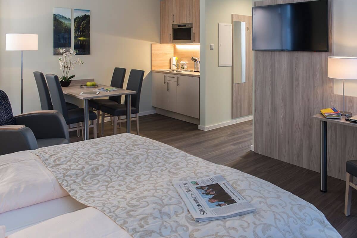 Una habitación de hotel con una cama, una zona de comedor con sillas y una mesa, un área de cocina con armarios y un microondas, y un televisor montado en la pared.