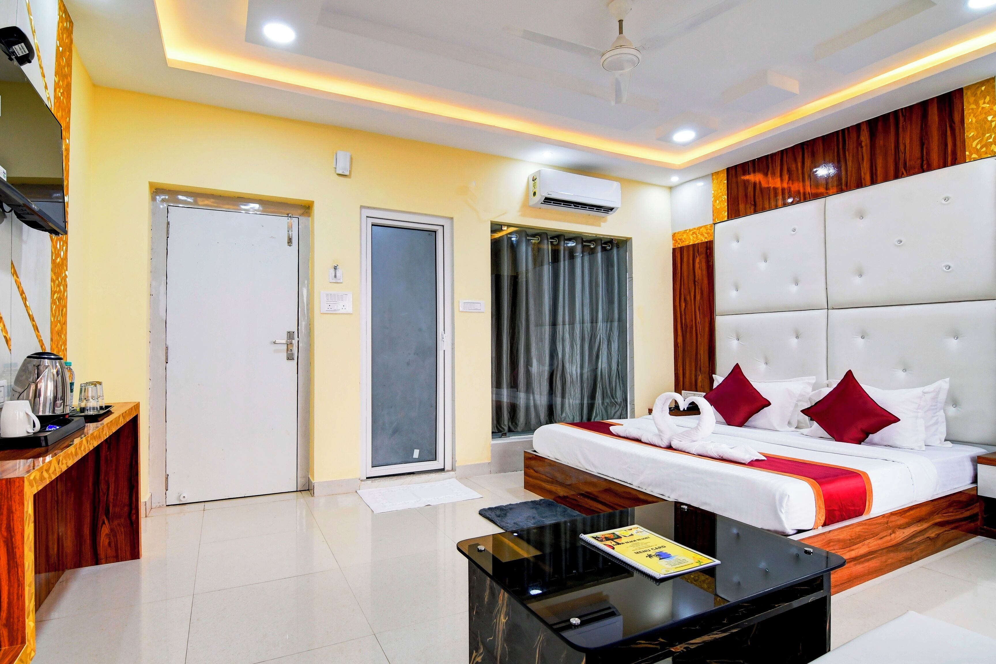 A hotel room with a bed, a coffee table, a desk with a kettle, and an air conditioning unit.