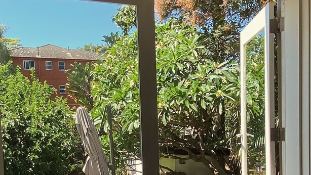 Vista a través de una puerta corrediza de vidrio hacia un patio exterior con mobiliario de madera y jardín.