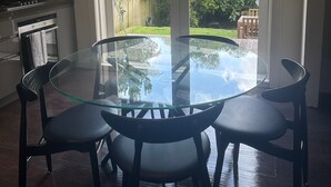 Un comedor con una mesa de vidrio y cuatro sillas, una cocina con estufa y alacenas, y un puerta corrediza de vidrio que da a un patio trasero con vegetación.