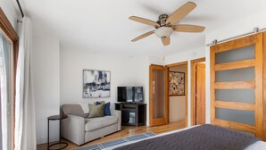 Quarto com cama, ventilador de teto, televisão, sofá e porta com painéis de vidro jateado.