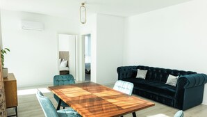 A modern living room with a wooden dining table, four blue upholstered chairs, a dark blue sofa with white cushions, and a wooden cabinet.