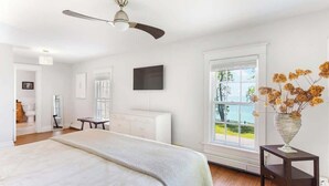 Un dormitorio con cama, ventilador de techo, ventana con vista al exterior y un florero con flores sobre una mesita de noche.