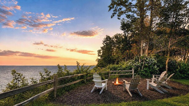 Una terraza con sillas Adirondack, una fogata y vista al océano al atardecer.